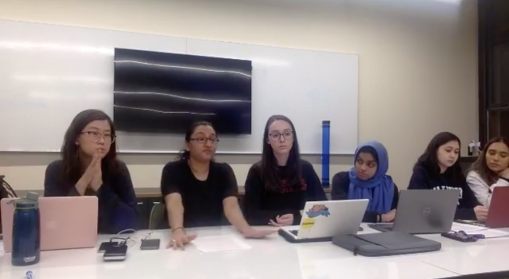 Six members of undergraduate student government sitting at a table with laptops in front of them. A TV and a whiteboard are behind them.